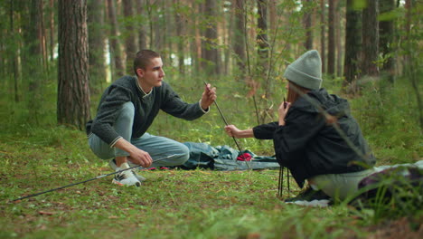lovers setting up tent during forest camping as woman hands tent poles to man while he looks at her affectionately under soft natural light surrounded by trees and peaceful outdoor environment