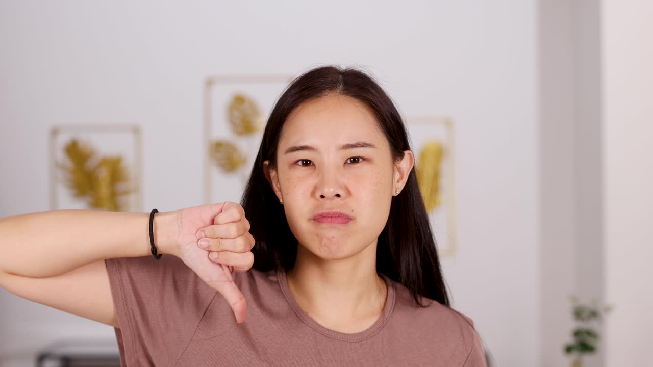 Young woman in casual shirt shows disappointment with thumbs down, neutral lighting, indoor setting