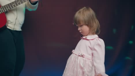 una niña con un vestido vintage baila en el escenario, su padre toca la guitarra acústica
