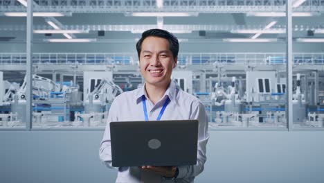 Asian Male Professional Worker Standing With His Laptop At Factory Digitalization, He Is Looking At The Camera With A Smile