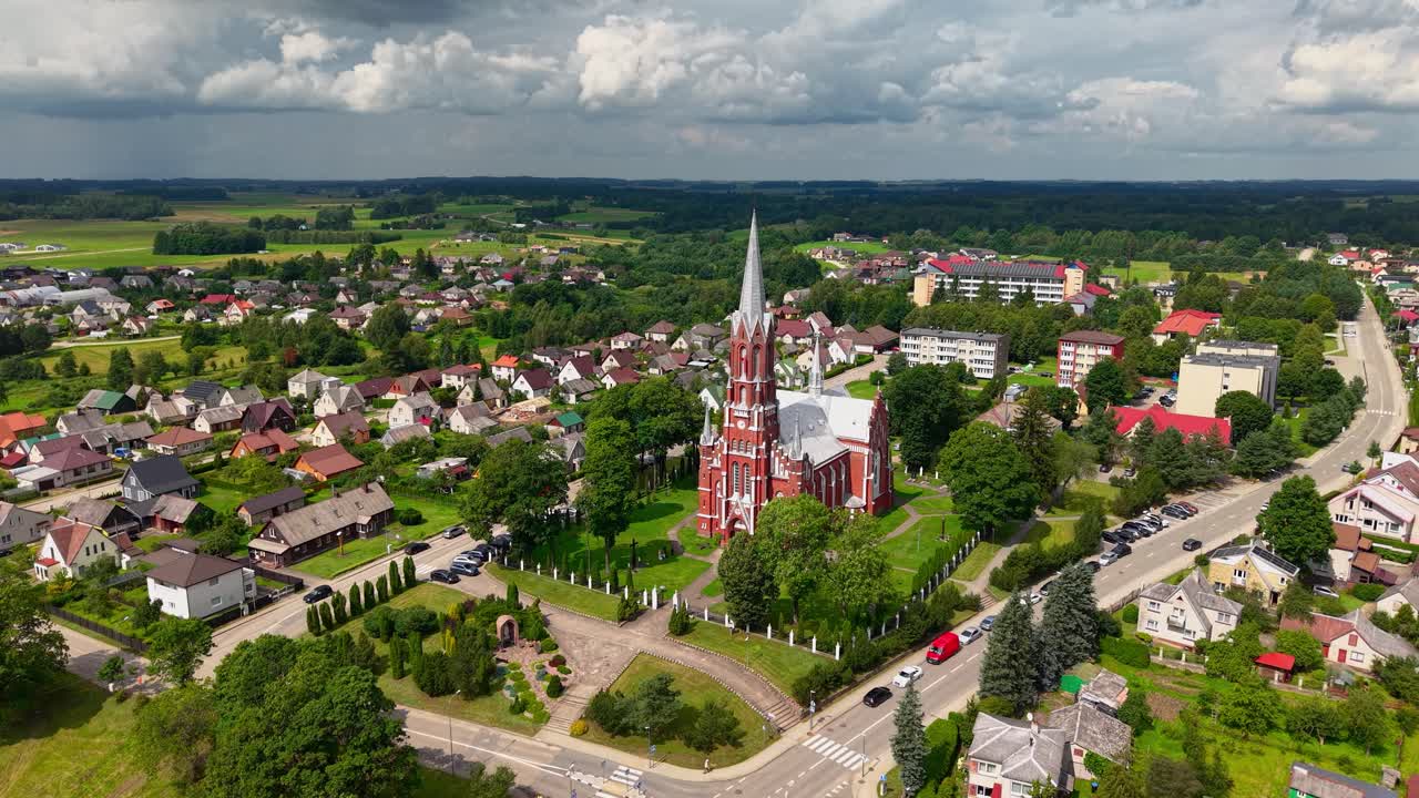 Scenic aerial view of Šilalė St. Francis of Assisi Church surrounded by colorful homes, lush greenery, and winding roads under a dramatic summer sky in Lithuania