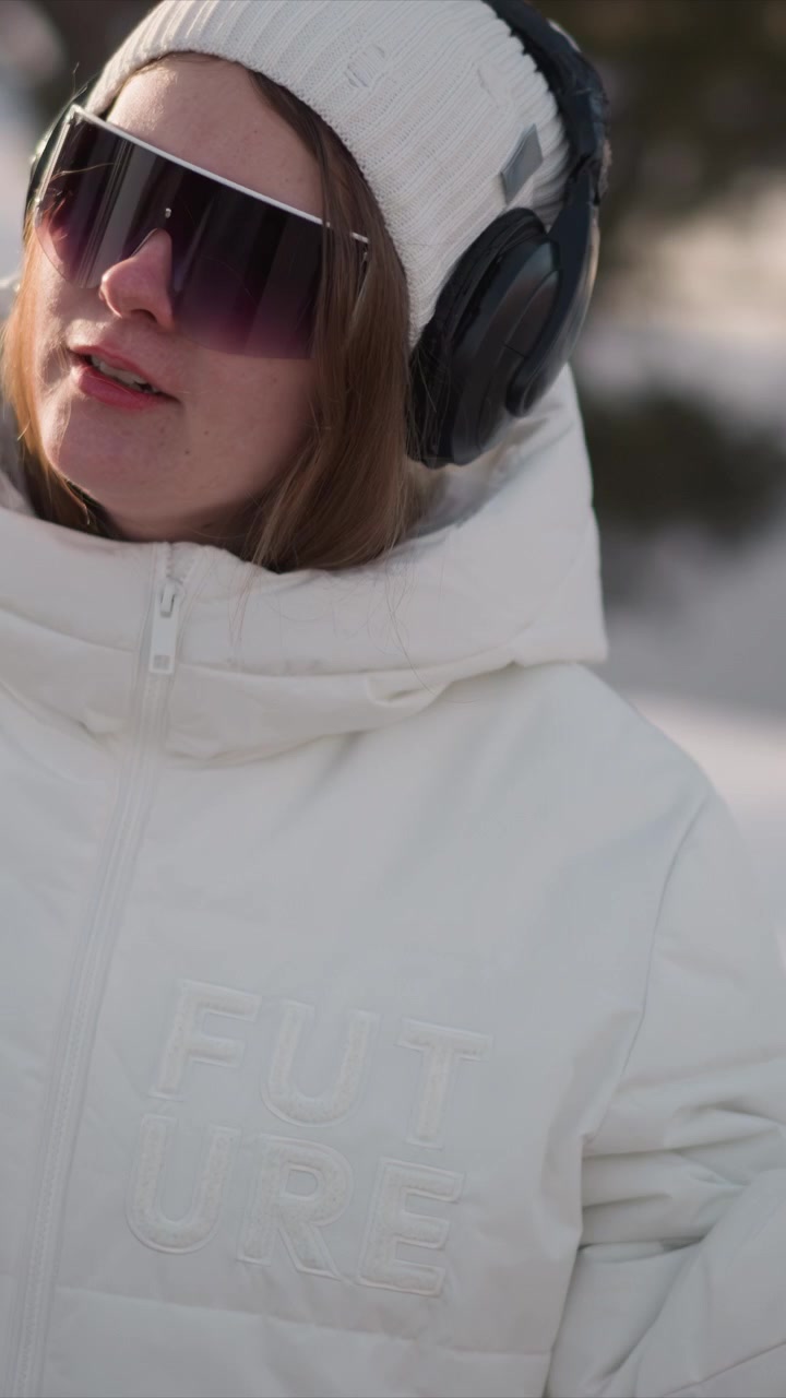 Woman wearing white puffer jacket and knit hat uses her smartphone outdoors in cold weather. Woman has large black headphones over her head visible nail polish on hands and word FUTURE