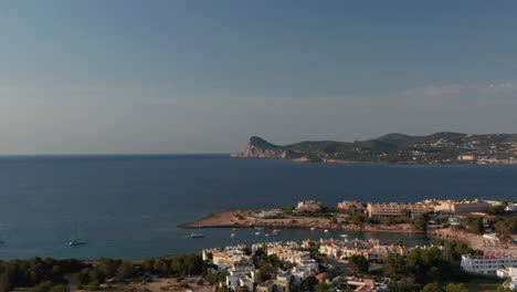 Establisher-shot-of-early-morning-view-of-silent-and-calm-sea-with-boats-and-yachts-parked-surrounded-by-mountain-and-beautiful-houses-with-greenery-around-in-Ibiza-in-Spain