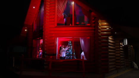 cozy atmosphere before christmas holidays. family decorating christmas tree together. couple with children prepare for winter celebration. view from outdoors.