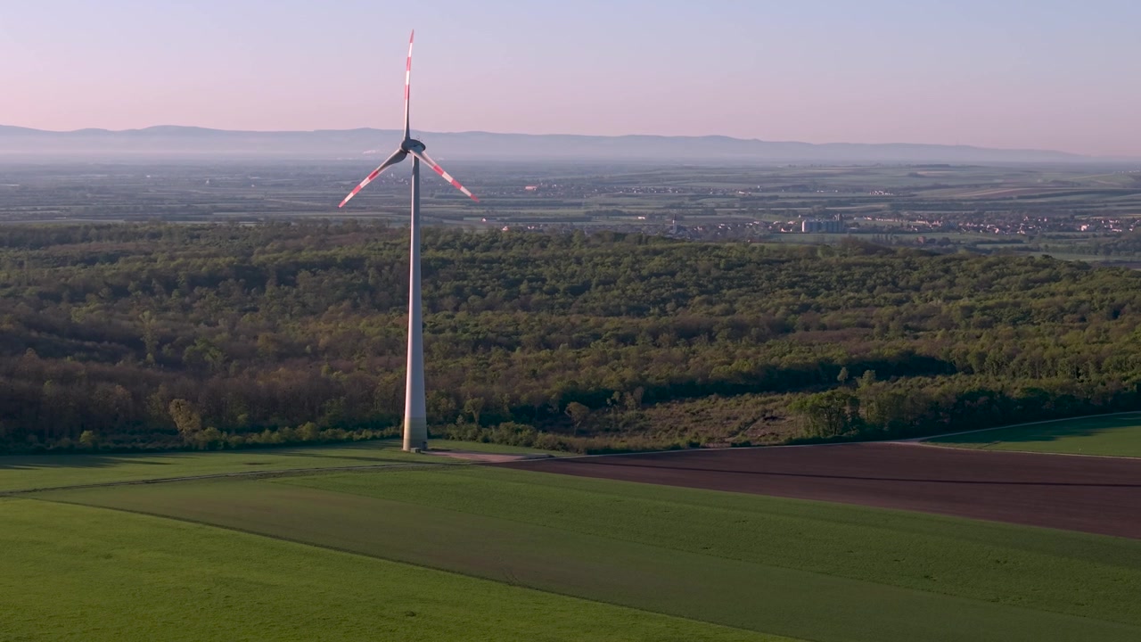 Vista aerea di una singola turbina eolica all'alba su un paesaggio agricolo. Turbina eolica bianca con marcature rosse che si erge sopra campi verdi e marroni. Infrastruttura di energia rinnovabile alla luce del mattino