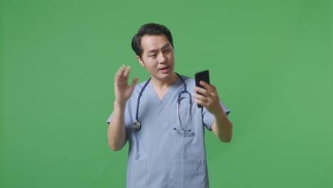 스테토스코프를 들고 있는 아시아 남성 의사 가 손을 흔들고 병원 에서 녹색 화면 배경 에 서 있는 동안 스마트폰 으로 비디오 통화를 하고 있다