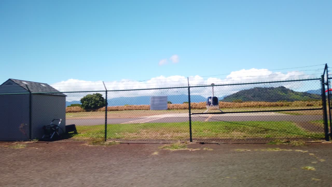 Gimbal wide shot from a moving vehicle approaching a chain link fence in front of a small helicopter at a heliport on the island of Kaua'i in Hawai'i