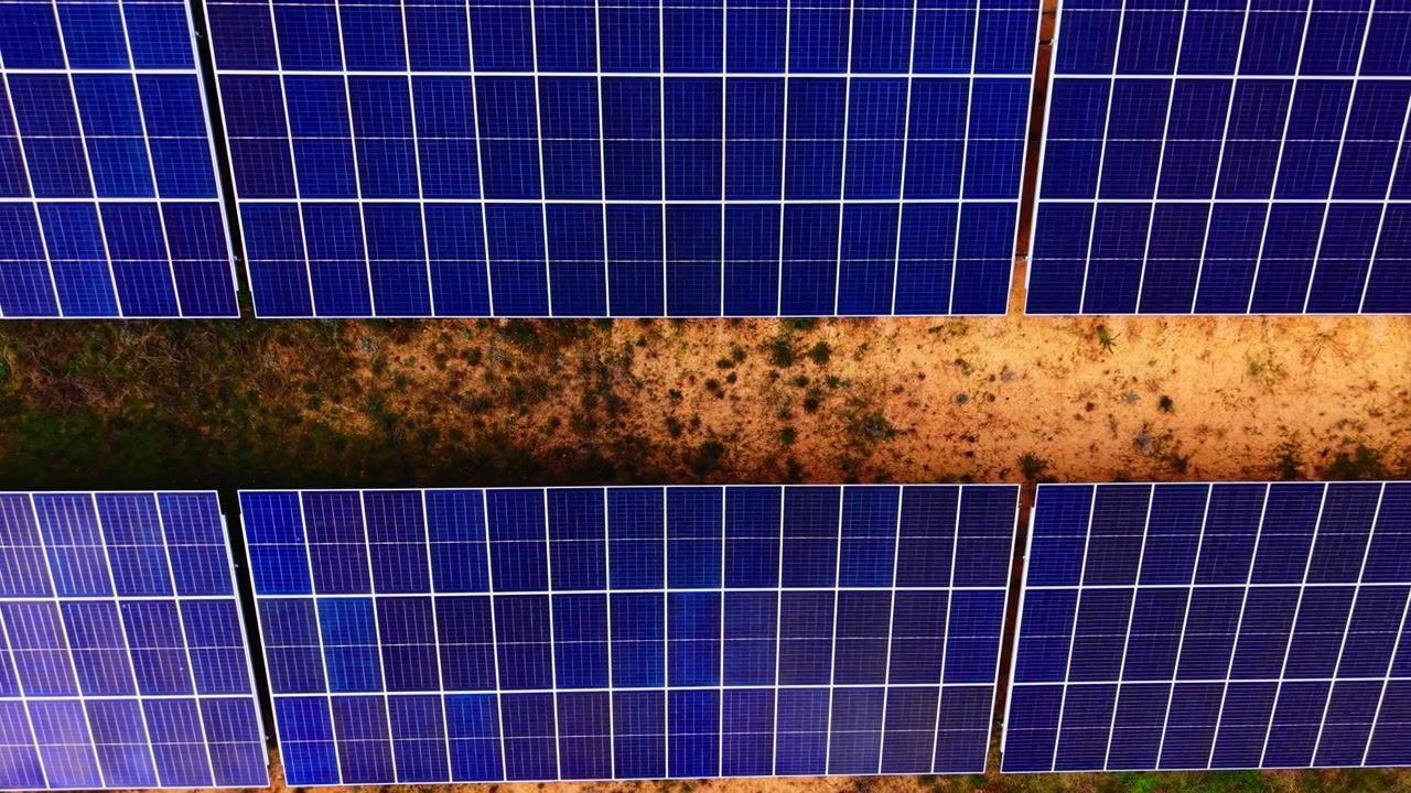 File di pannelli solari divise da un sentiero sterrato. Vista aerea da drone di moduli fotovoltaici blu scuro incorniciati da terreno nudo e vegetazione verde irregolare
