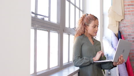 Biracial-female-fashion-designer-standing-by-window-and-using-laptop-and-in-studio,-slow-motion