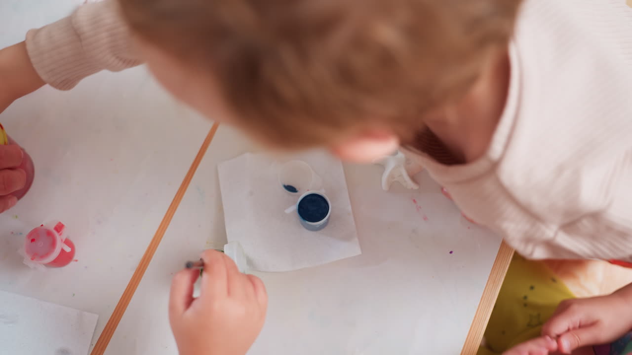 Hand view of instructor holding small blue paint container and opening lid while assisting child during art activity, preparing supplies for painting session with children in classroom setting
