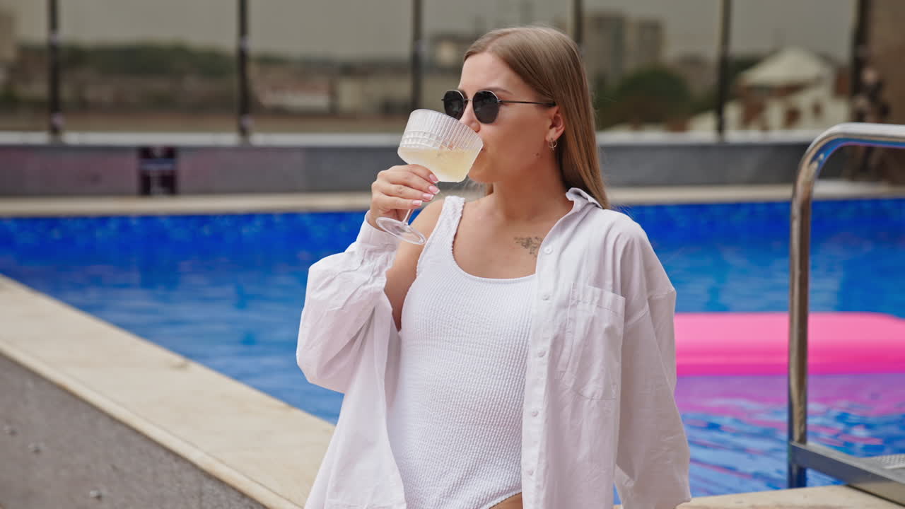 Relaxed calm lady in white shirt drinking cocktail near the swimming pool. Girl enjoying leisure time near the pool.
