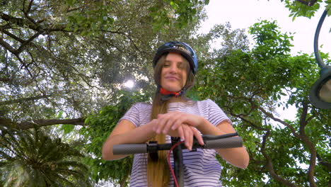 vista desde abajo de una joven divertida con scooter eléctrico y casco que muestra la lengua a la cámara