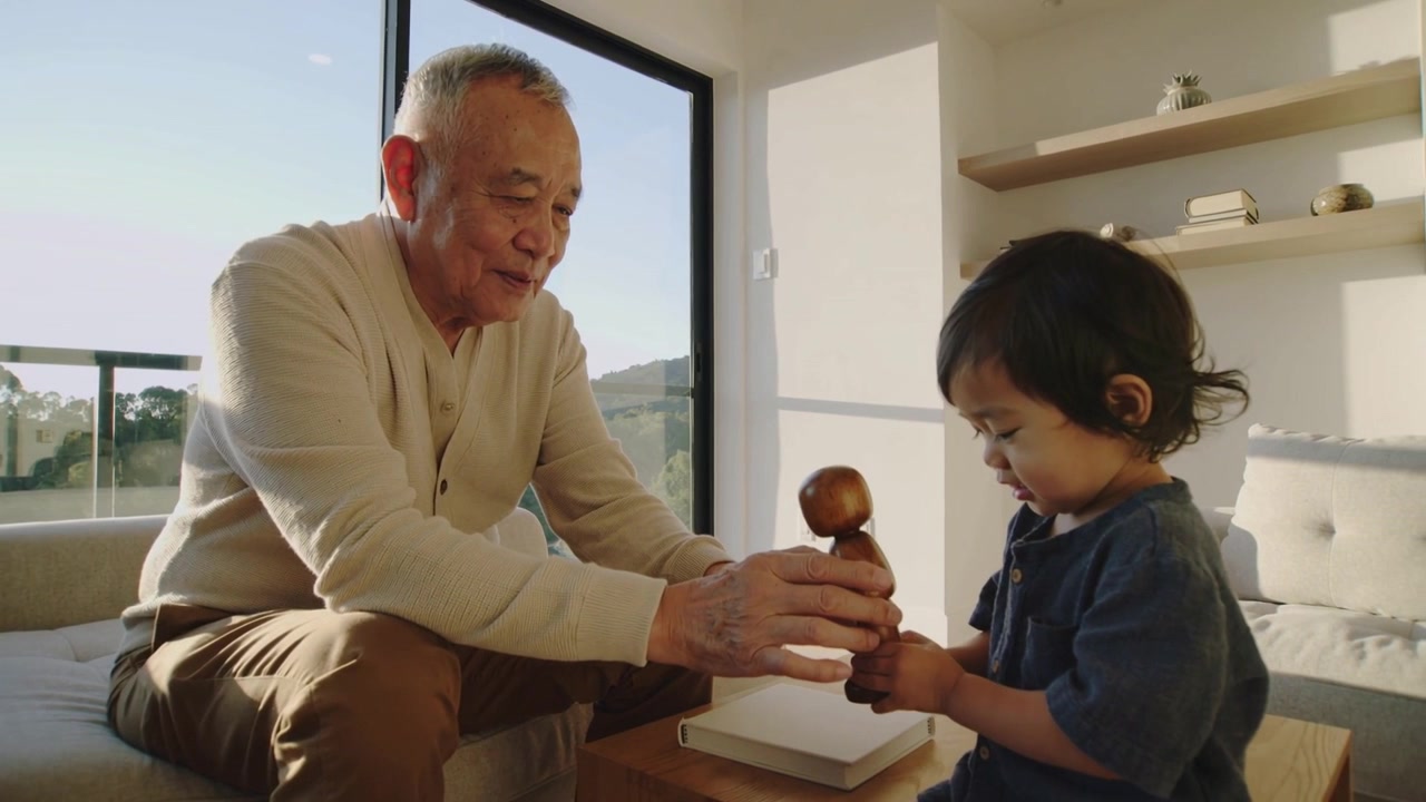 自宅でベージュのセーターを着た年配の女性が青いトップスを着た幼児を導き、木のおもちゃの持ち方を教えている