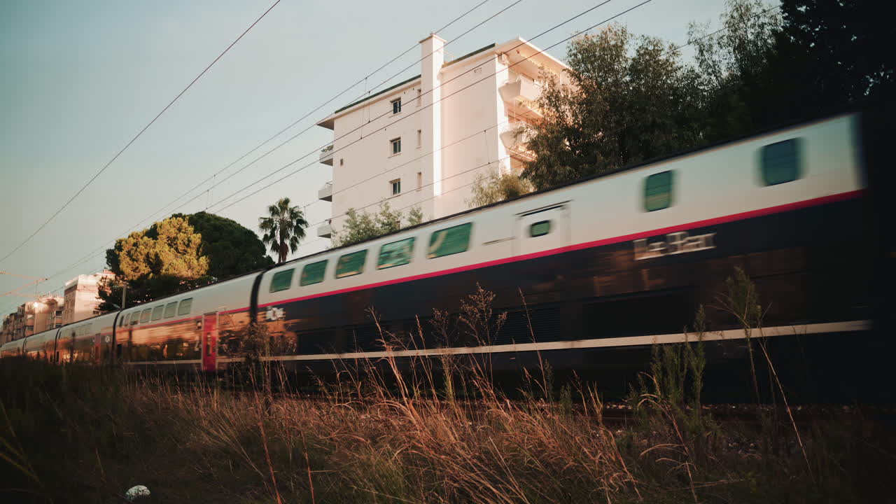 A blue commuter train speeds past a residential building, creating strong motion blur against the still architecture
