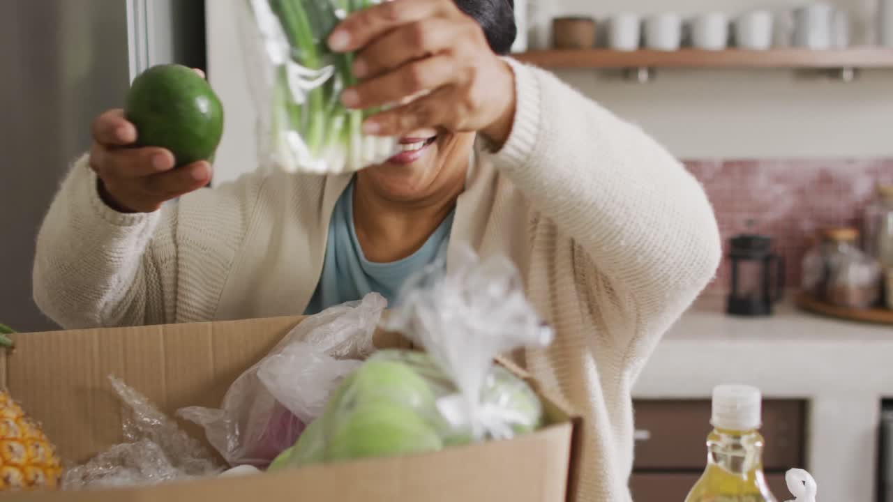 mujer biracial mayor sonriente agarrando verduras de la caja en la cocina sola