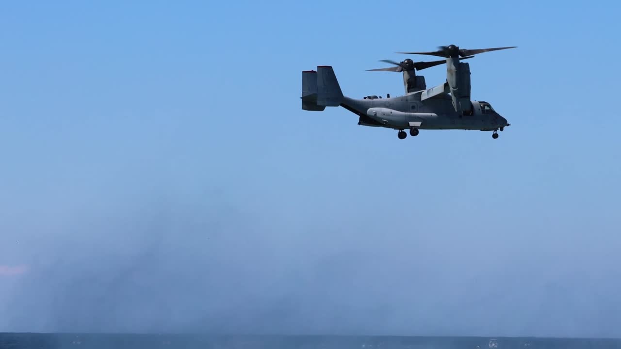 A tiltrotor aircraft hovers above the ocean, creating a visible spray against a clear blue sky.