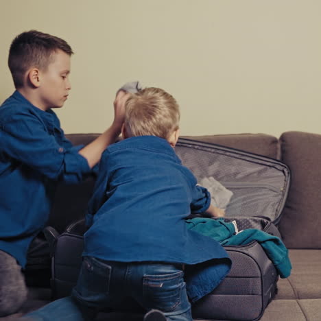 two curious boys are putting clothes into the suitcase for a trip at home. active brothers packing the things in the travelling bag together in the room. slow motion.