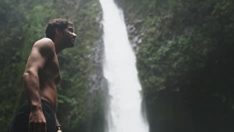 low angle of shirtless man looking up at rushing waterfall in green rain forest