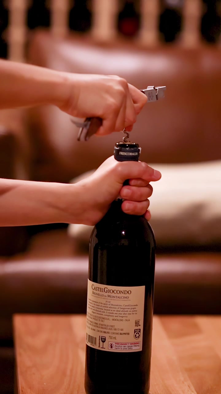 Una persona usa un sacacorchos para abrir una botella de vino sobre una mesa de madera en una bodega de vinos cálidamente iluminada, con un suave desenfoque de fondo.