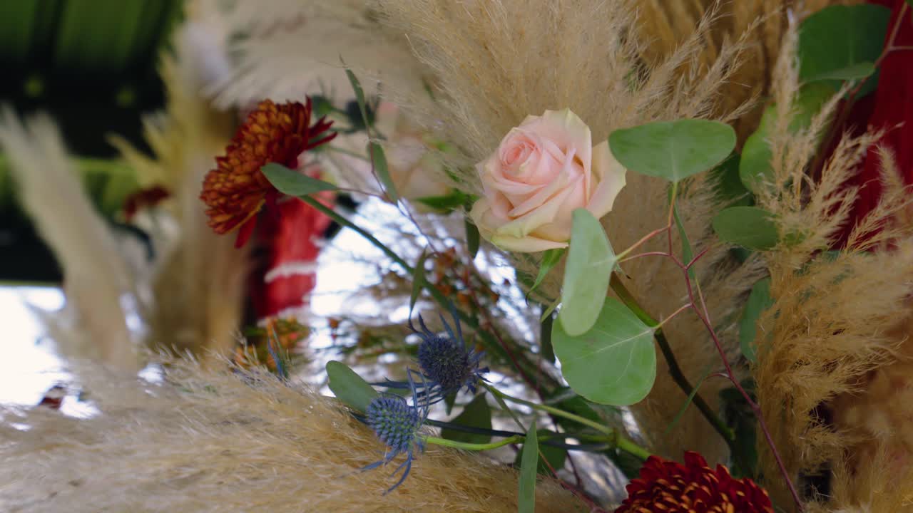 Close-up of a pink rose gently swaying among pampas grass and fall florals on a wedding arch, capturing the natural movement and romantic outdoor setting.