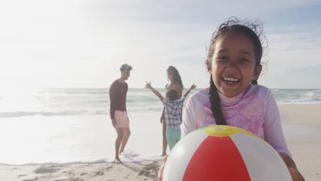 happy hispanic girl holding ball on beach with family in background