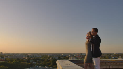 Ein-Reizendes,-Süßes,-Glückliches-Paar-Sitzt-Auf-Dem-Dach-Mit-Herrlichem-Blick-Auf-Den-Sonnenuntergang-über-Der-Großstadt.-Sie-Lieben-Sich-Und-Umarmen-Sich-Sehr-Zärtlich,-Die-Sonne-Geht-Zwischen-Ihre-Haare