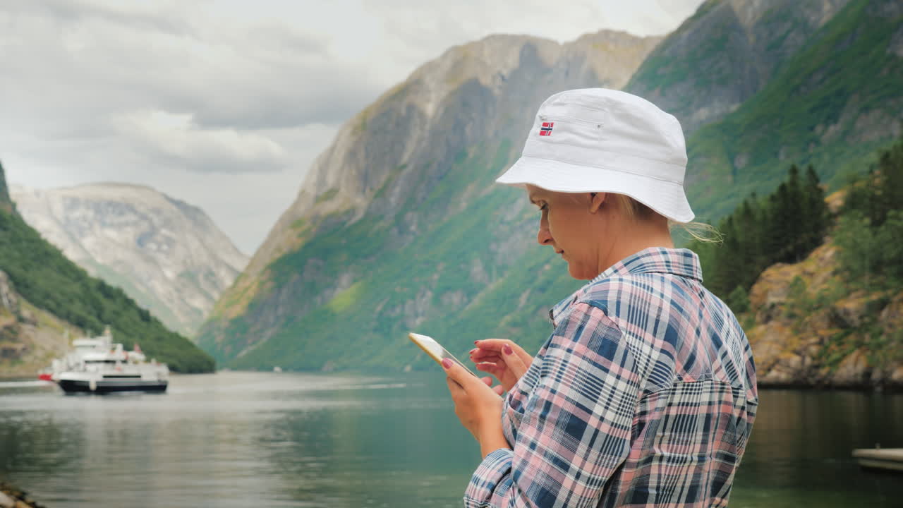 한 관광객이 멀리 있는 그림 같은 노르웨이 피요르드를 배경으로 스마트폰을 사용합니다.