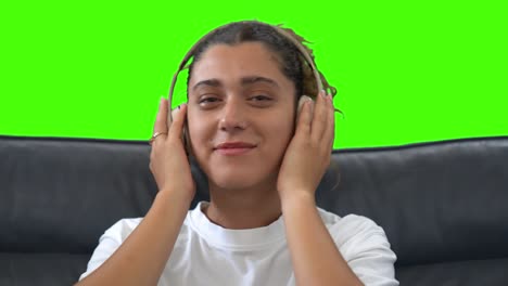 Girl-Putting-On-Wireless-Headphones,-Sitting-On-Couch,-Green-Screen-Background