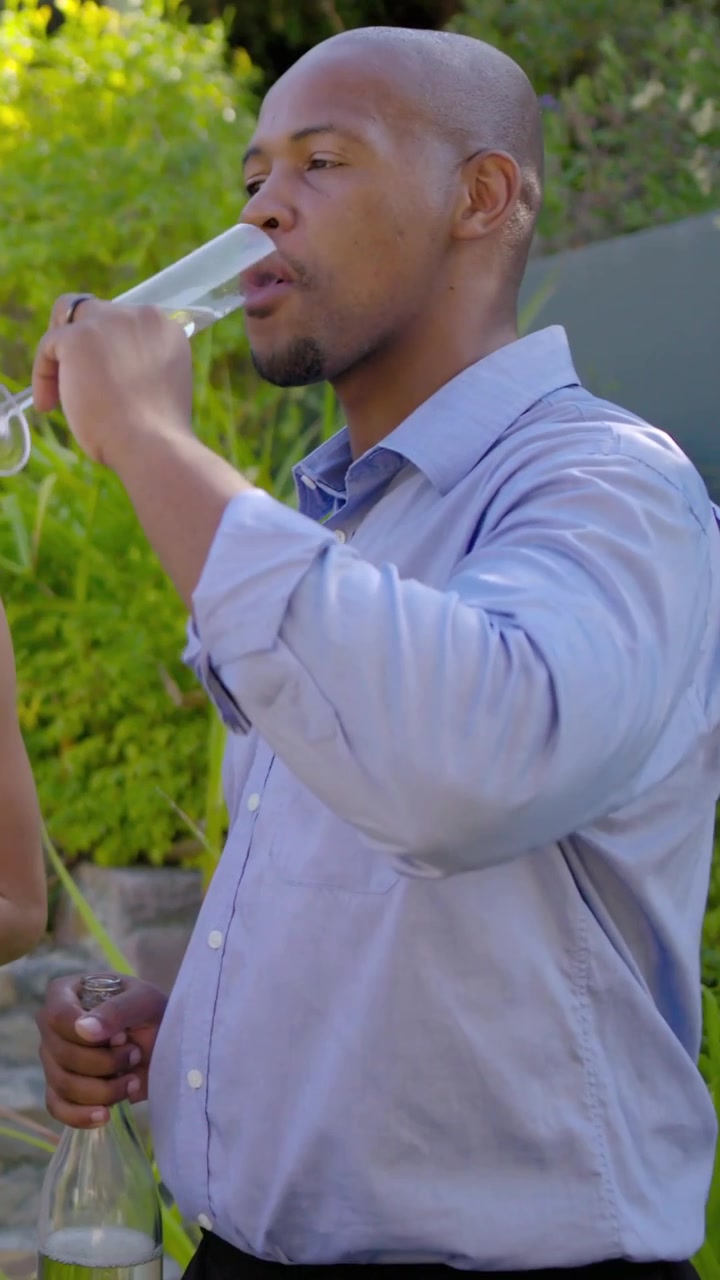 Vertikales Video: Lächelnder Mann im blauen Hemd erhebt ein Glas, trinkt und hält dabei eine Karaffe im Garten.