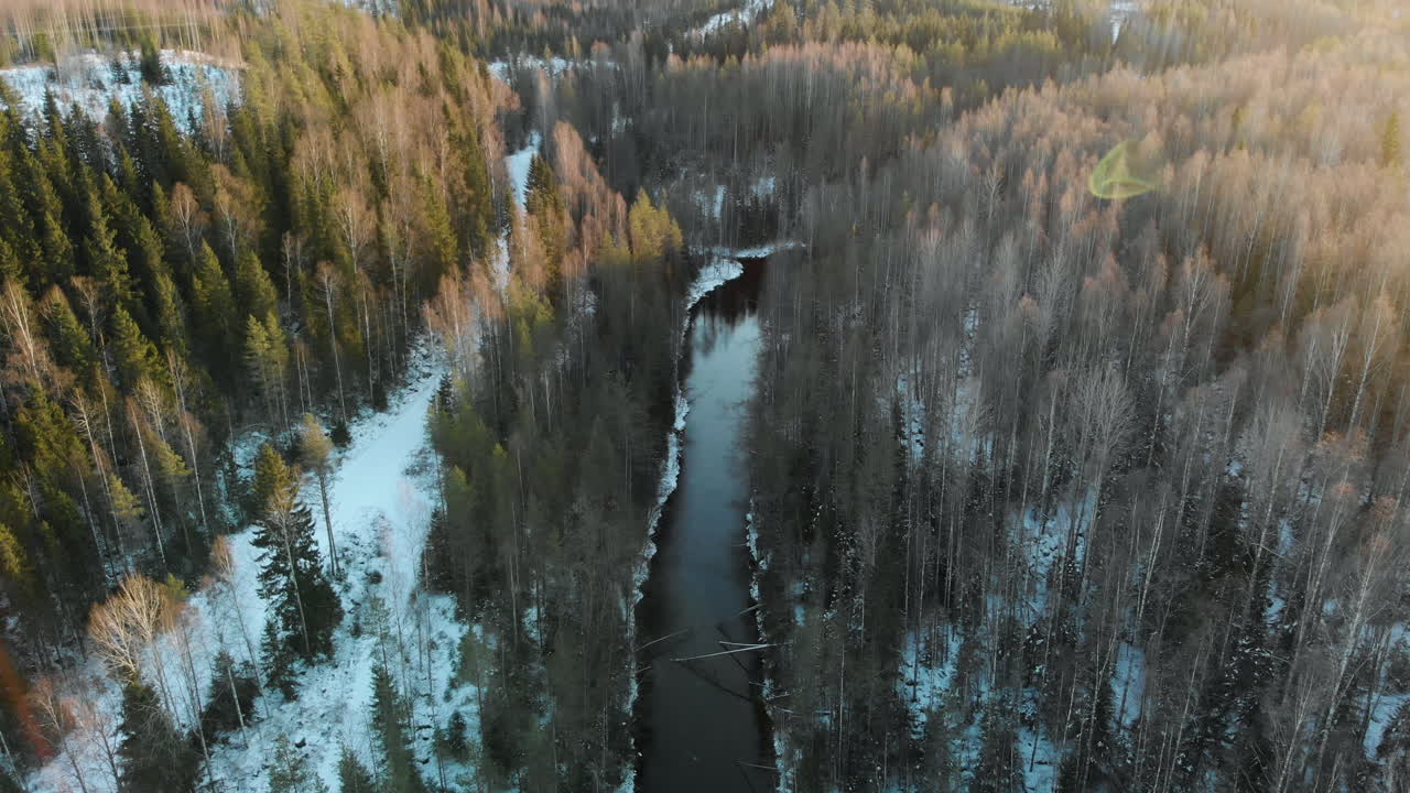 Aerial, rising, tilt up, drone shot, sun flares, above a river, surrounded by leafless forest and first snow on the ground, on a sunny winter day, near Joensuu, North Karelia, Finland