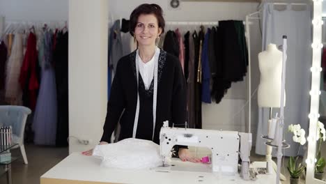 Portrait-of-beautiful-young-short-haired-woman-seamstress-in-elegant-black-sweater-standing-in-workplace,-measuring-tape-on-neck,-looking-and-smiling-at-camera.-Dummy,-garments,-sketches-and-sewing-machine-are-visible.Slow-motion