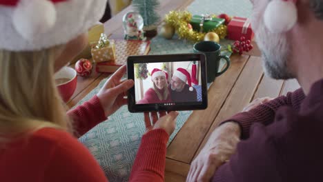 Lächelndes-Kaukasisches-Paar-Mit-Weihnachtsmützen,-Das-Ein-Tablet-Für-Einen-Weihnachtsvideoanruf-Mit-Einem-Paar-Auf-Dem-Bildschirm-Verwendet