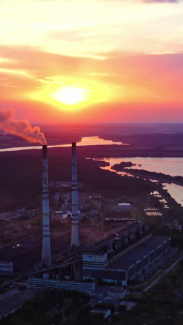 factory on colorful sky background at sunset. harmful smoke filling the air from industrial pipe in the evening. manufacturing near the river. aerial view. vertical video