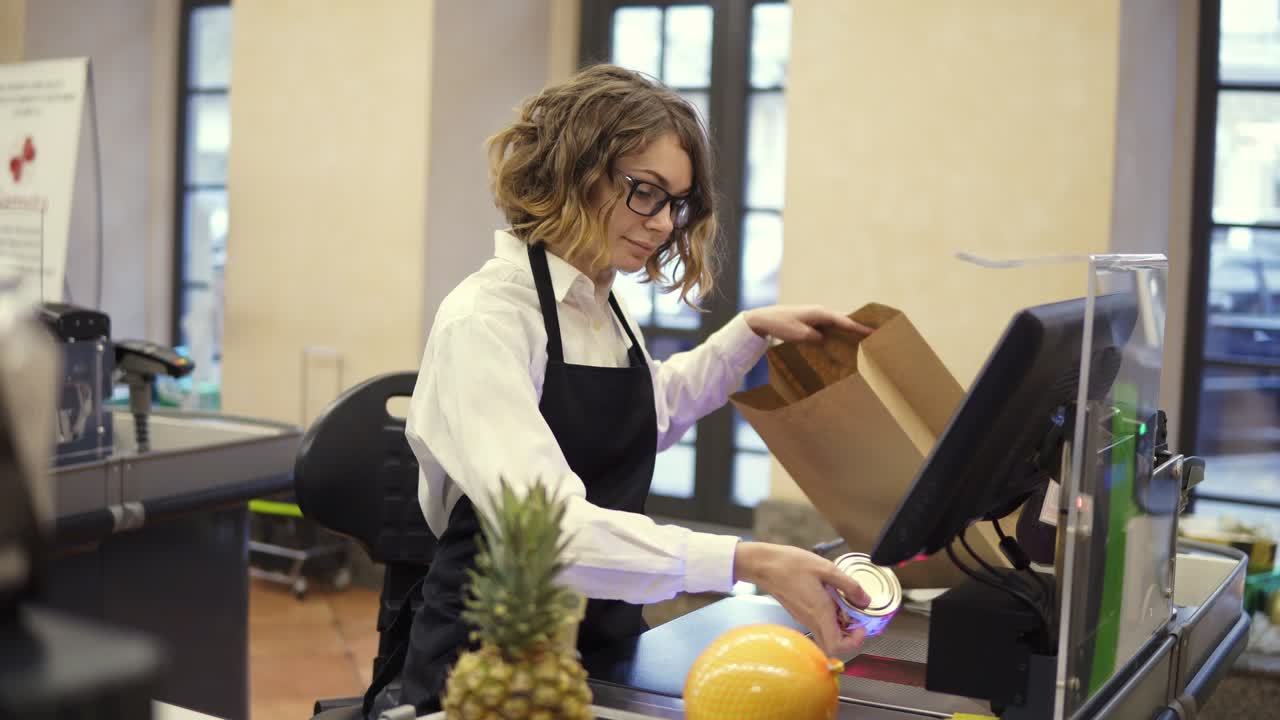 Cheerful slender saleswoman in white shirt and black apron scanning product, fruits at checkout counter in bright supermarket and putting it into brown paper bag