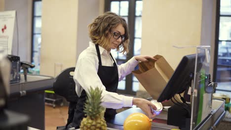 mujer de ventas delgada y alegre en camisa blanca y delantal negro escaneando producto, frutas en el mostrador de caja en un supermercado brillante y poniéndolo en una bolsa de papel marrón