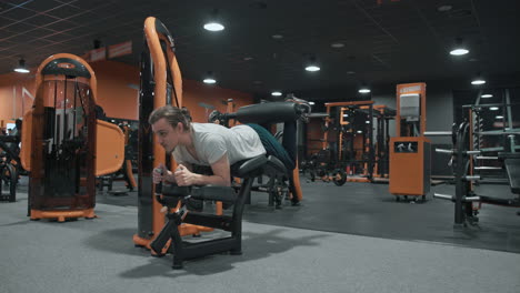 wide shot of 20s man training legs curls on machine in indoor fitness club gym