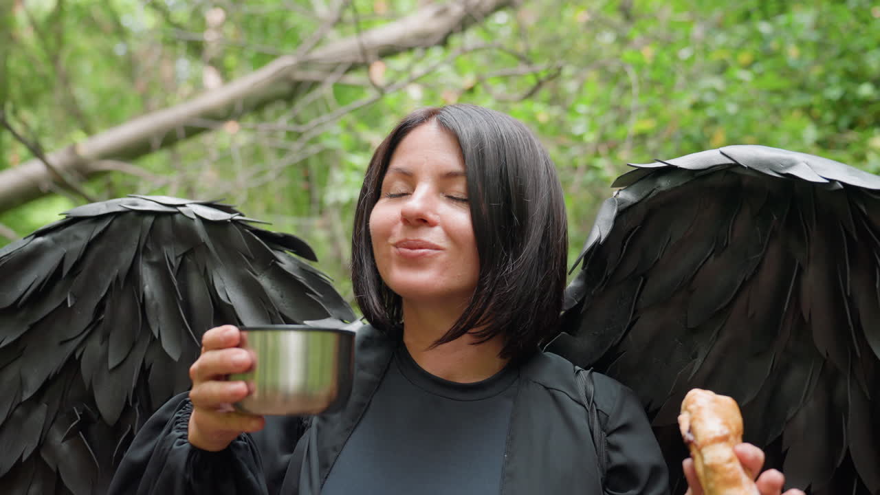 Dark fairy dressed in black costume with feathered wings sips drink from metal cup while holding sausage roll in hand, surrounded by lush forest greenery, expressing calm