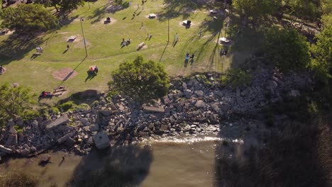 aerial drone shot of people relaxing and resting in green grass park at shore of river plate at sunset