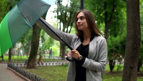 hermosa mujer comprobando si está lloviendo o no y luego abriendo su colorido paraguas de pie en el parque de la ciudad. disparo en cámara lenta