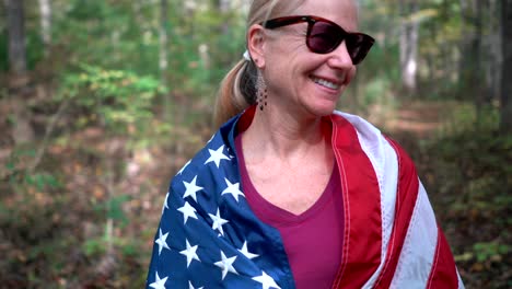 Kamera-Dreht-Sich-Um-Eine-Hübsche,-Blonde-Frau-In-Einem-Wald,-Um-Die-Sich-Eine-Amerikanische-Flagge-Gewickelt-Hat