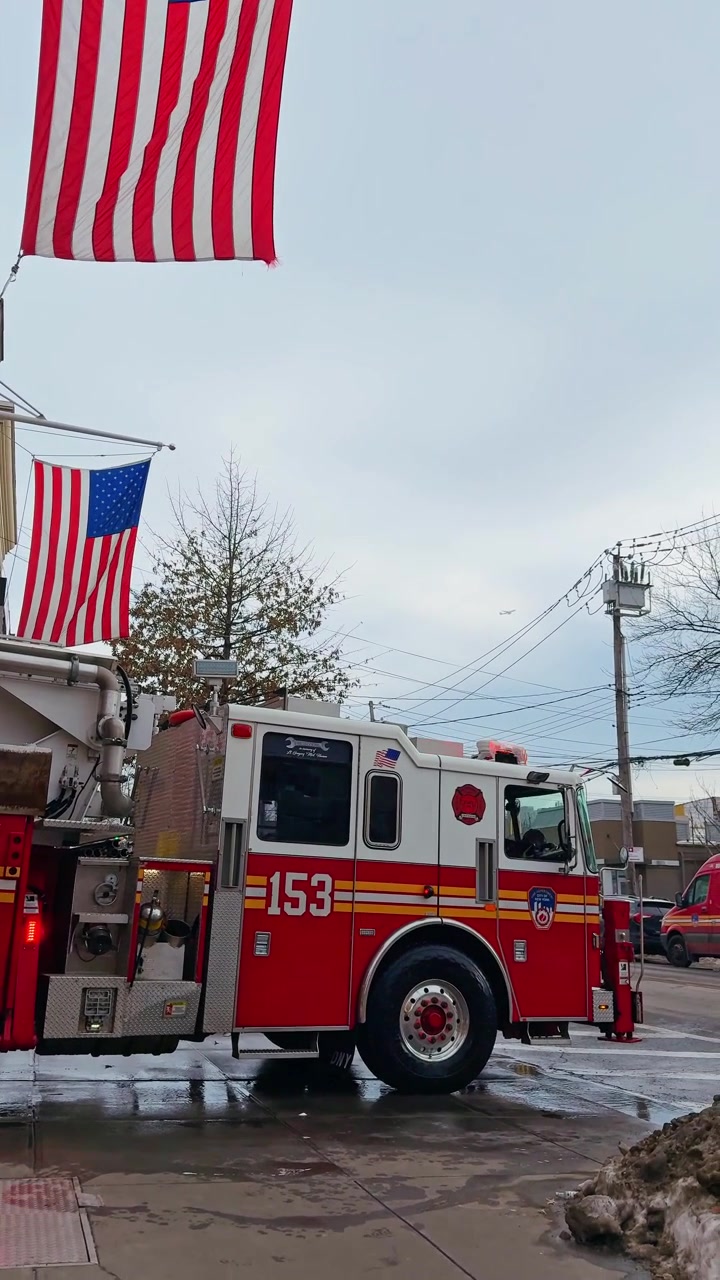 New York, ABD, 25 Ocak 2026: Şehir sokağında kırmızı ve beyaz itfaiye aracının yan profili. Şehir manzaralı, Amerikan bayrağı altında ıslak bir köşeye park etmiş 153 numaralı itfaiye aracının profili.