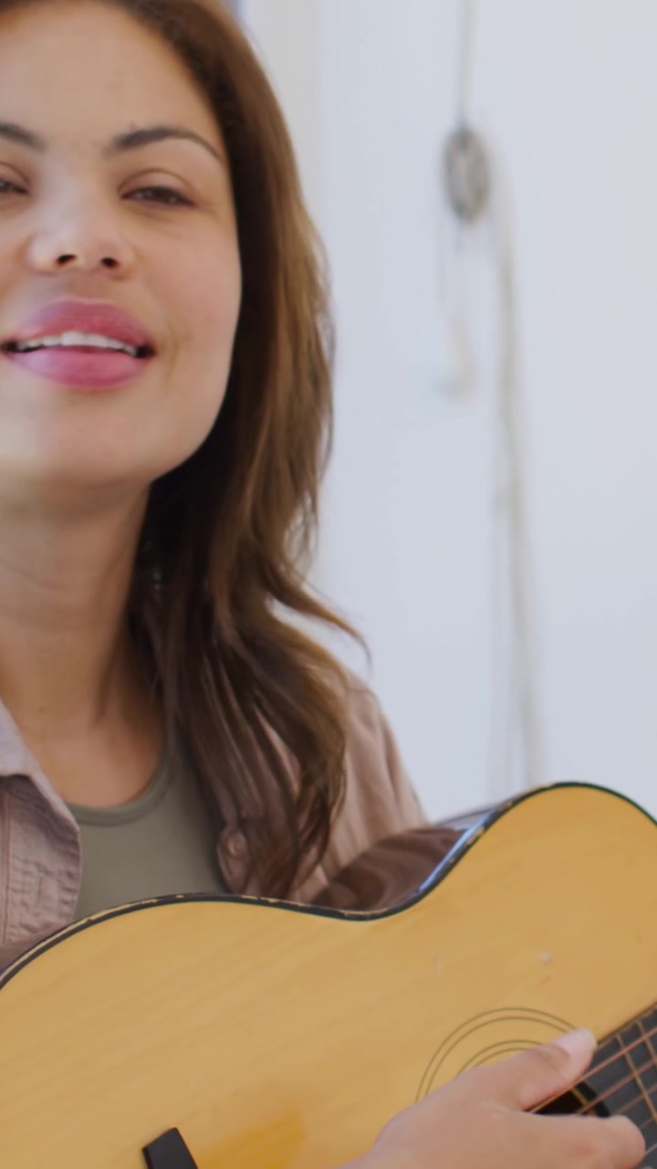 Video verticale: Cantante che canticchia, strimpella e canta, esercitandosi a casa in camicia chiara, con chitarra e orologio.