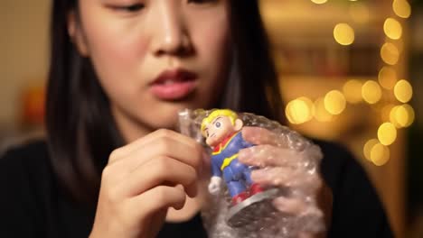 a young woman carefully unwraps a bubble-wrapped toy with a focused expression, surrounded by beautiful bokeh lights in the background