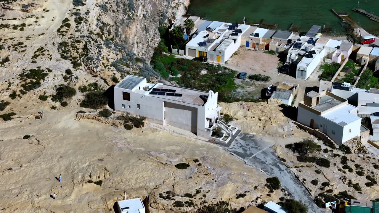 Letecký pohled dronem na moderní bílou minimalistickou budovu se solárními panely zasazenou do drsných vápencových útesů zálivu Dwejra na Gozu na Maltě pod jasnou světlou letní oblohou s tyrkysovou vodou