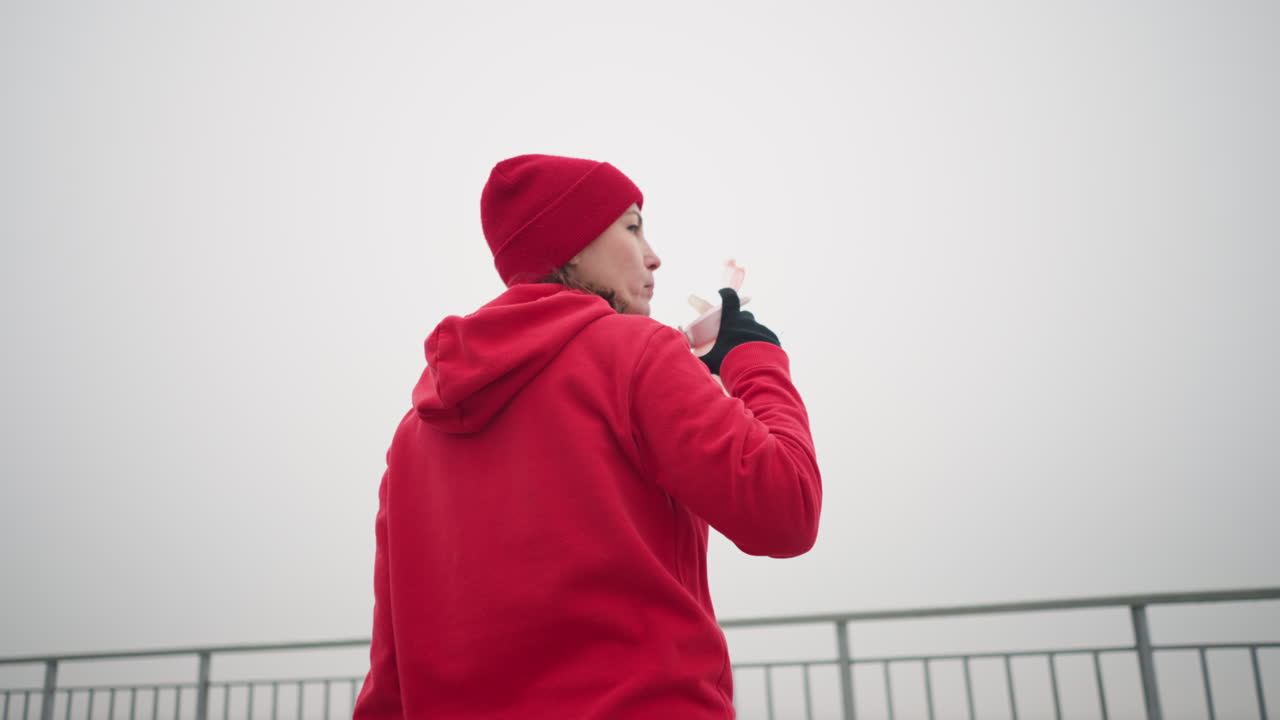 vista trasera de una mujer con capucha roja y gorra bebiendo agua al aire libre usando una botella rosa que cae, en una atmósfera de niebla, con un fondo borroso de barandilla de hierro y un paisaje brumoso
