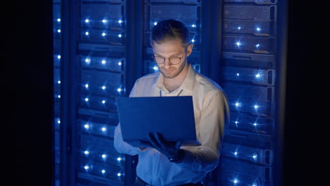 male network engineer doing a system check standing in the server room with his laptop. at data center men server specialists inspecting working system and hardware of rack server computer cabinets