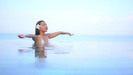 Una-Mujer-Joven,-Con-El-Agua-A-La-Altura-Del-Pecho-En-Una-Piscina-De-Borde-Infinito,-Levanta-Los-Brazos-Del-Agua-Hacia-El-Cielo-Y-Sonríe