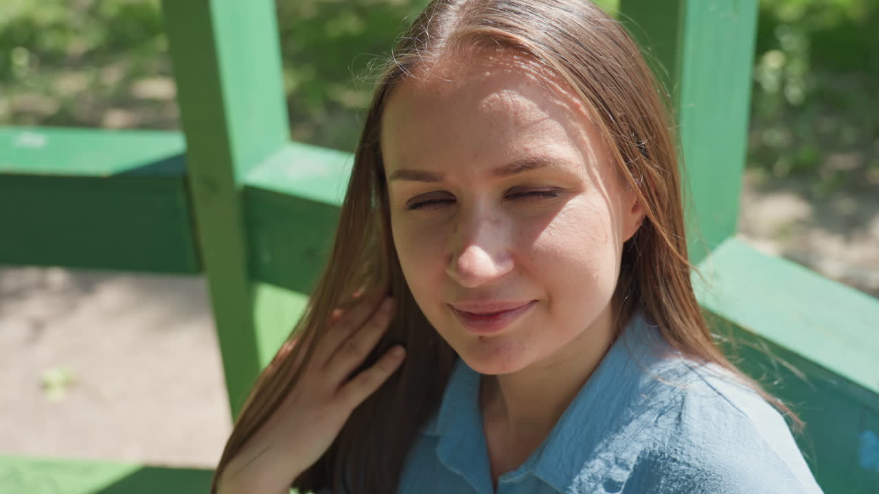 Mujer informal disfrutando de un día soleado al aire libre; modelo tranquila sentada en la naturaleza con la luz del sol iluminando su rostro; mujer caucásica en calma que parece relajada mientras está sentada en un banco bajo la luz natural del sol.