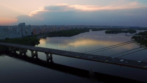 Drone-view-car-traffic-on-suspension-bridge-over-river-on-evening-city-landscape