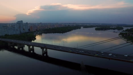 Drohnenansicht,-Autoverkehr-Auf-Der-Hängebrücke-über-Den-Fluss-In-Der-Abendlichen-Stadtlandschaft